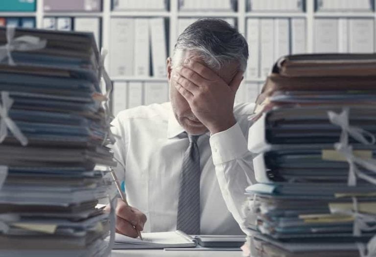 A man in a shirt and tie sits at a desk with his head in his hand, surrounded by large piles of files