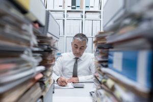 A man sits at a desk surrounded by files and piles of paper