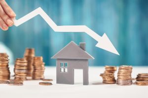 A small grey model house surrounded by piles of coins, with a woman's hand holding a white arrow pointing downwards above the house