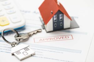 A small model house next to a key, with the word 'approved' stamped on a piece of paper