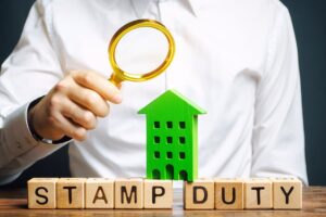 The words 'stamp duty' spelled out in wooden blocks, with a man holding a magnifying glass up to the letters