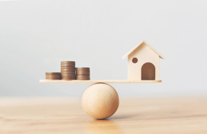 A basic wooden model of a home balanced with a pile of coins on top of a wooden ball