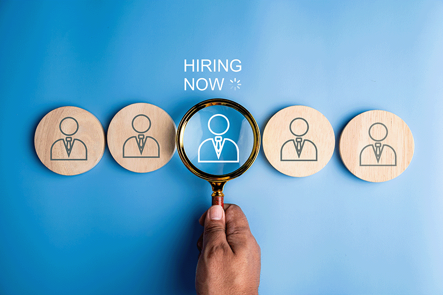 The words 'hiring now' above five discs with graphics of simple outlines of people. A magnifying glass is held over the central disc