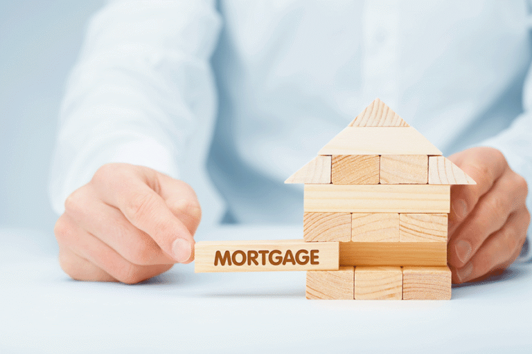 A wooden house made of small bricks, with one bearing the word 'mortgage' being slightly pulled out like the game of jenga