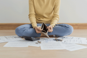 A woman sits cross-legged, surrounded by bills, holding open an empty wallet