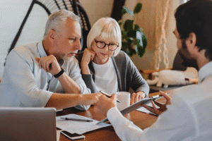 An older-looking couple with grey hair sign papers at a desk, assisted by a younger man
