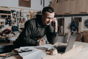 A man in a workshop surrounded by tools leans over a workstation containing an open laptop, while talking on the phone and taking notes.