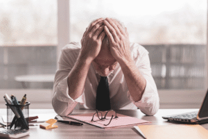 A man sits at an office desk with his head in his hands, looking tired and stressed