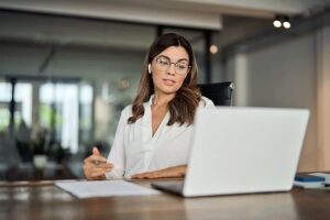 A woman sits at a desk in front of a laptop, taking notes