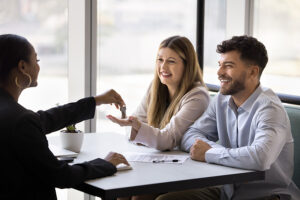 A happy couple are handed a set of house keys by a woman in a suit sitting at the other side of a desk