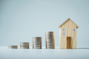 A small toy house is placed to the right of increasingly large piles of coins