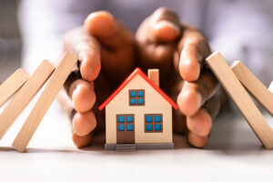 Close up of hands surrounding a small wooden house, preventing dominos from falling into it