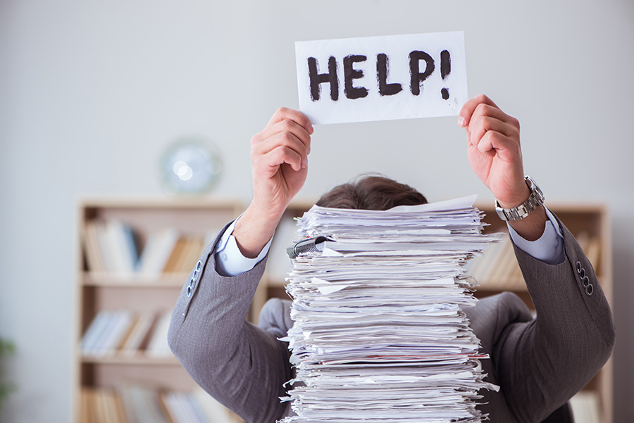 A man at a desk hidden behind a large pile of papers, holding up a piece of paper with 'help' written on it