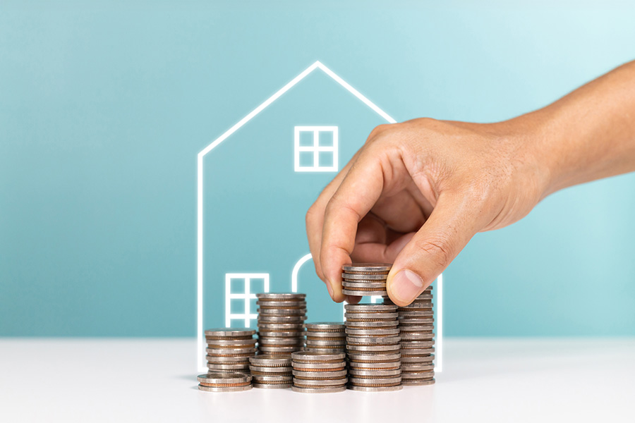 An outline of a house, with pile of coins in front of it and a hand placing more coins on the pile