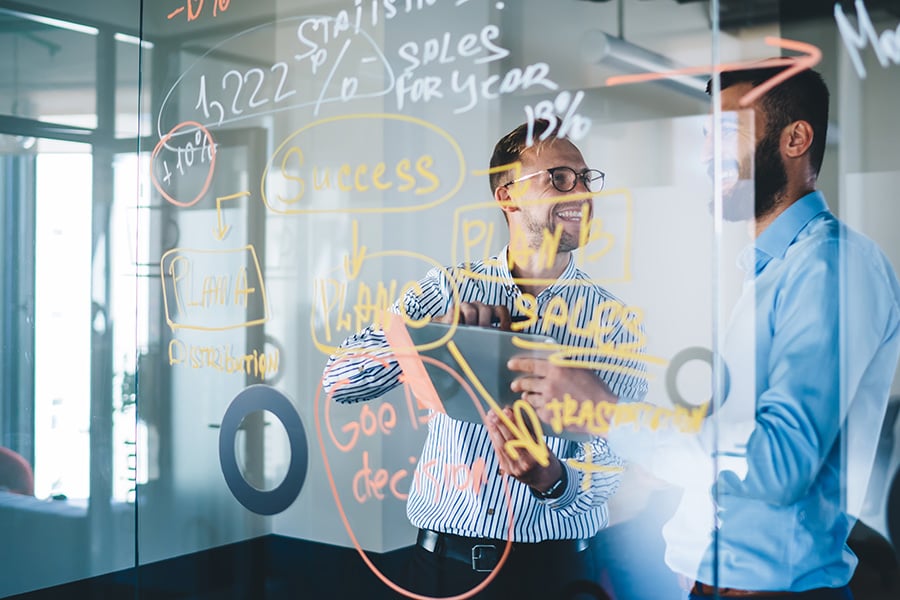 Two men write on a glass ideas board in coloured pen