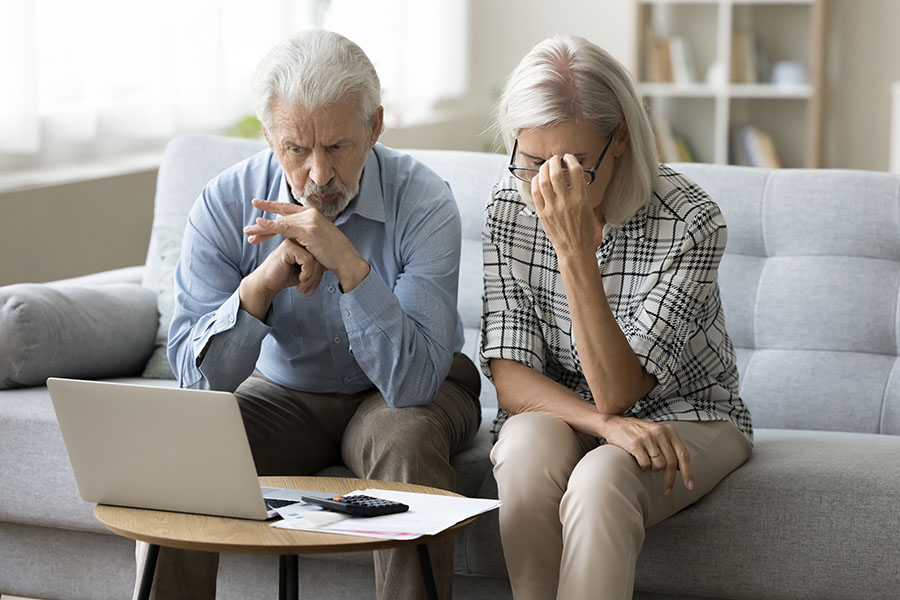 An older couple sitting on a sofa looking concerned, with a laptop and calculator in front of them