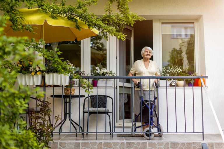 An older lady stands on the plant-filled balcony of a home