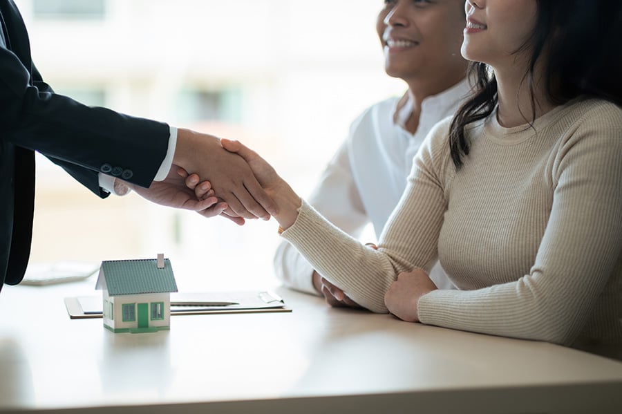 A smiling couple shake hands with a person just out of shot, with a small model of a house on the desk between them
