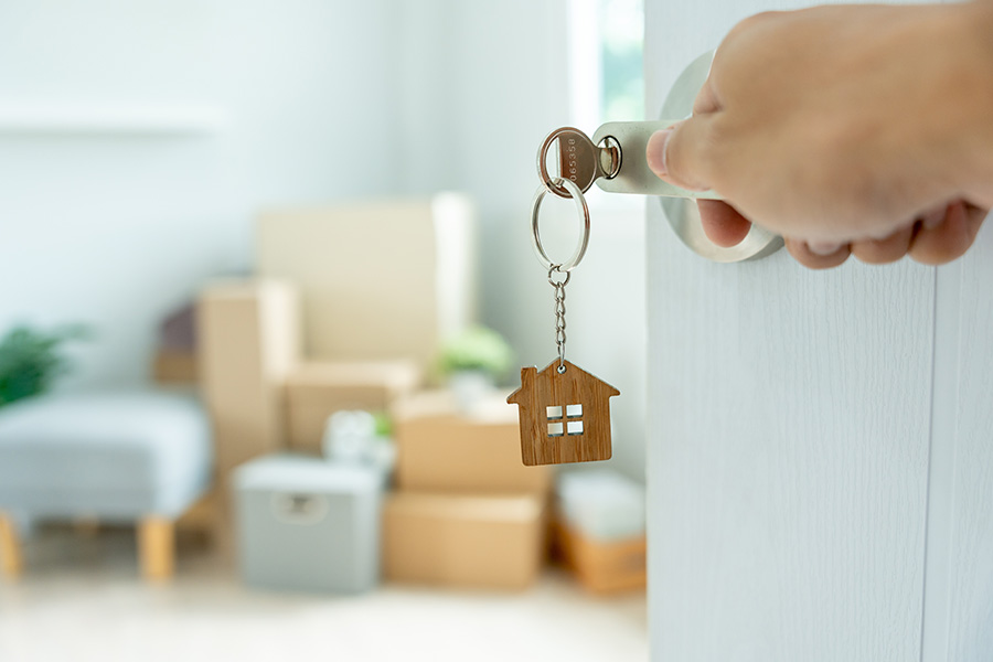 In the foreground a hand opens a door with a key in it, in the background are packing boxes and furniture, all in a pastel blue colour