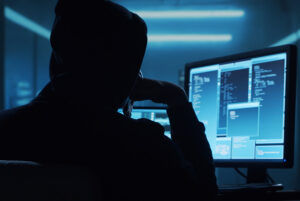 a hooded figure in shadows sits in front of a computer screen