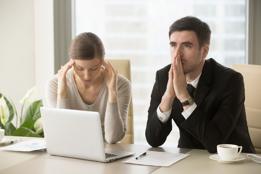 A man and woman sit at a desk with a laptop in front of them, both looking worried