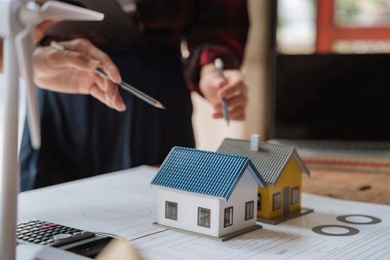 Two small model houses on top of papers on a desk, with two people in suits gesturing to the houses