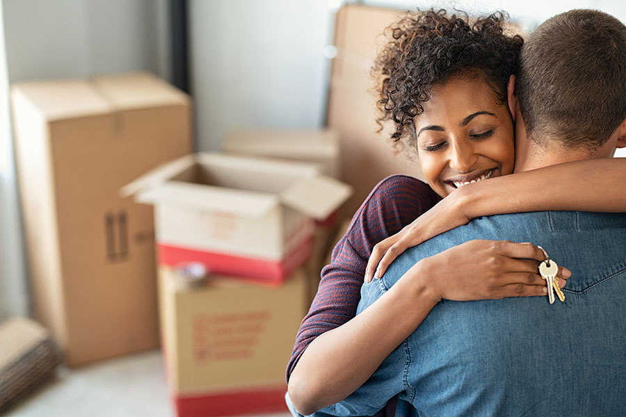 A couple hug holding keys to a home, in a room with packing boxes
