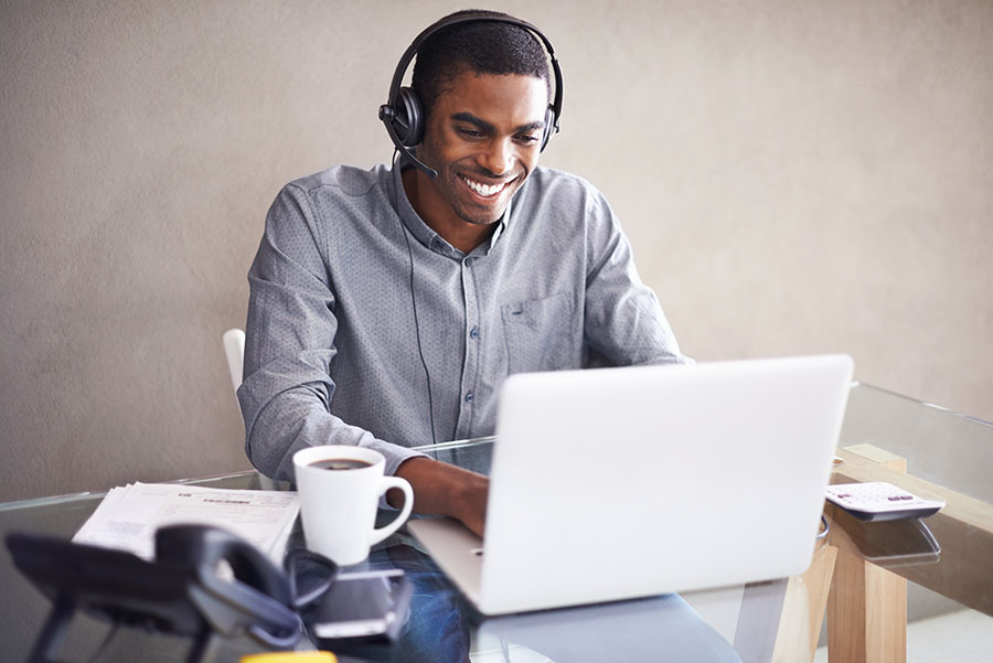 A man sits at a desk in front of an open laptop, wearing headphones and holding a pen