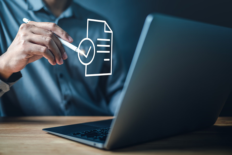 A close-up of an open laptop, with a person pointing to an overlaid graphic of a document with a tick mark.
