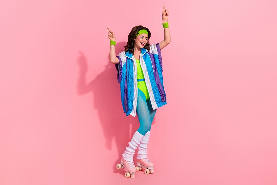 A woman wearing 80s-style lycra and roller boots in a dancing pose against a pink background