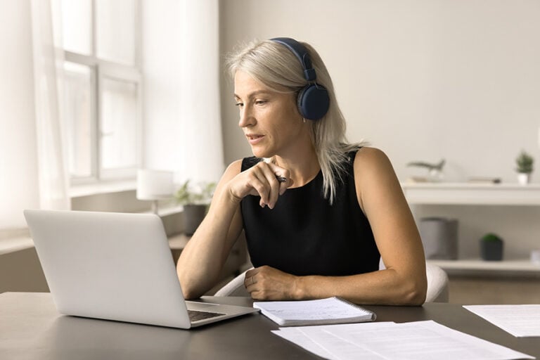 A woman sits at a desk looking at a laptop screen while wearing headphones