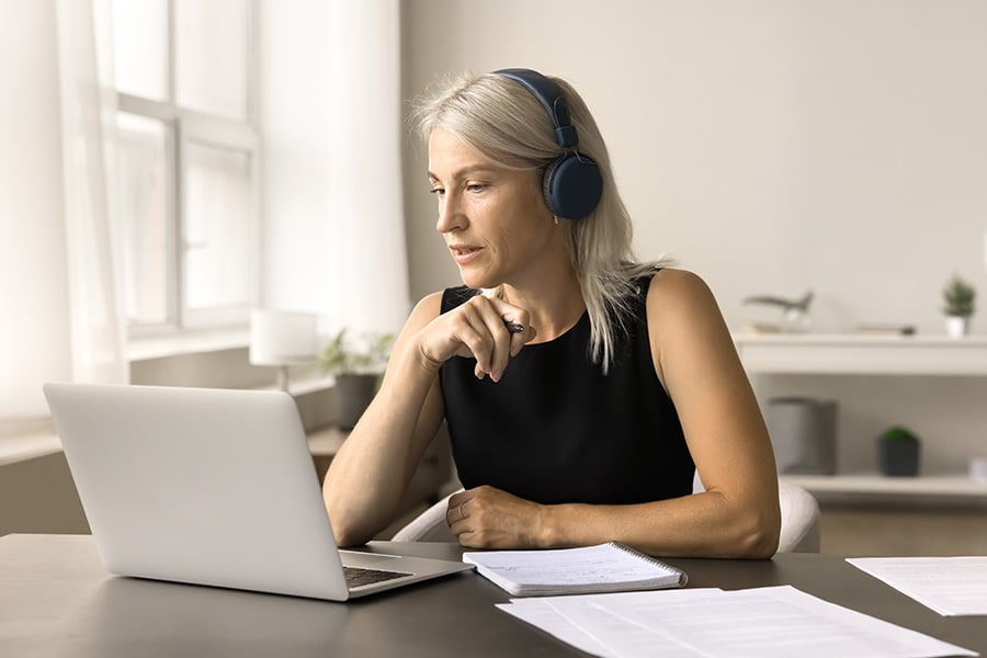 A woman sits at a desk looking at a laptop screen while wearing headphones