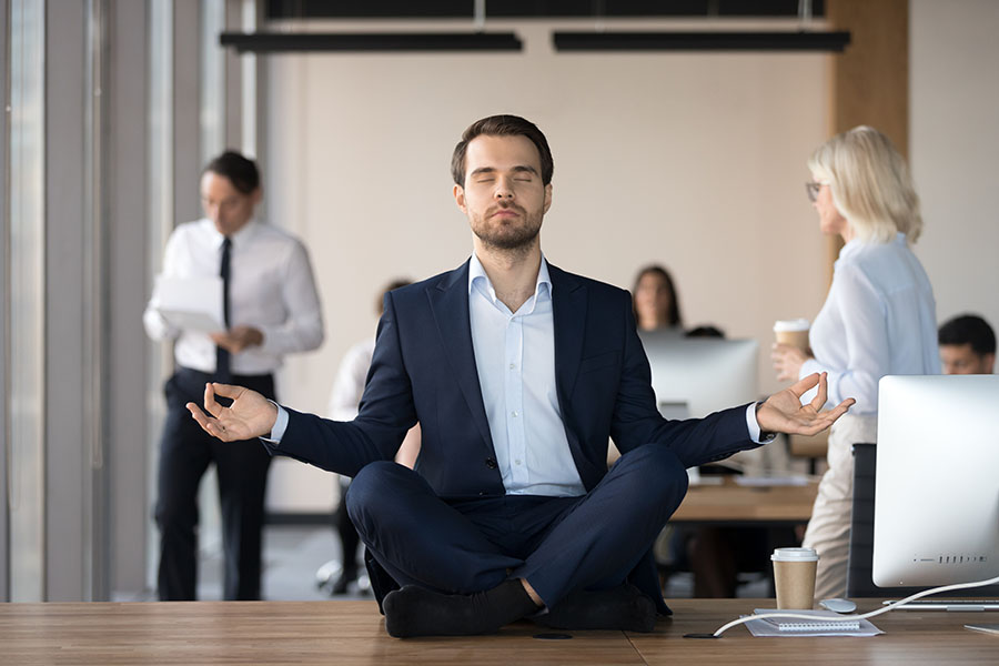 A man in a suit sits in a cross-legged yoga pose on a desk in an office