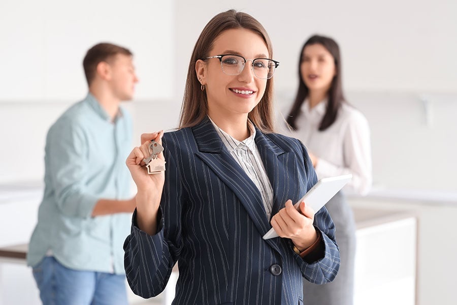 An estate agent holds up a key and a clipboard, with a couple in the background
