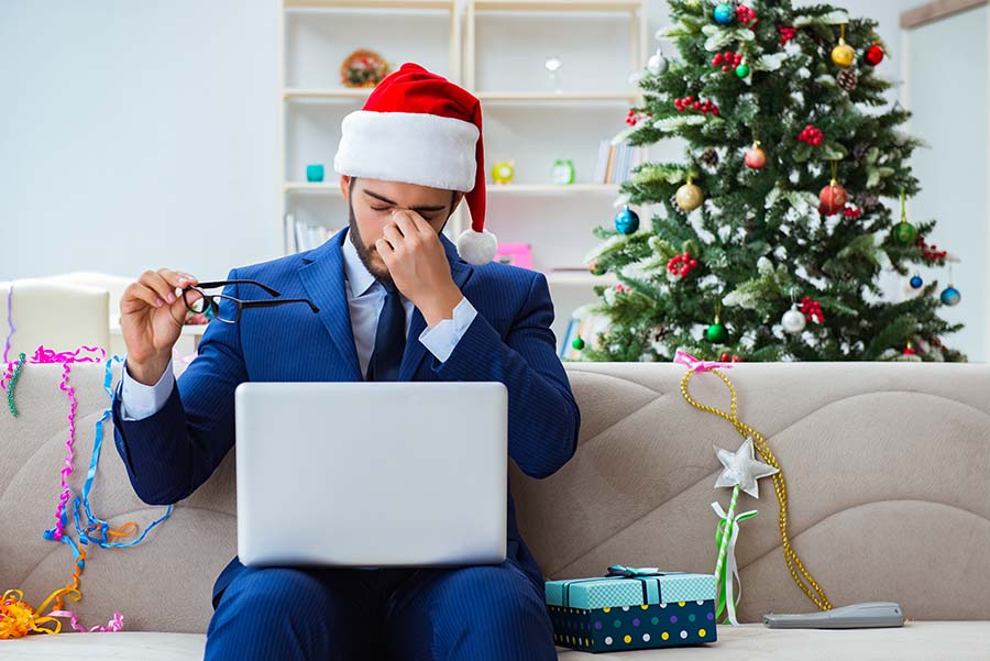 A man wearing a suit and santa hat sits on the sofa with a laptop, rubbing his eyes