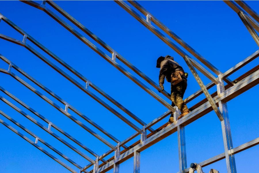 A construction worker balances on high up steel beams against a cloudless blue sky