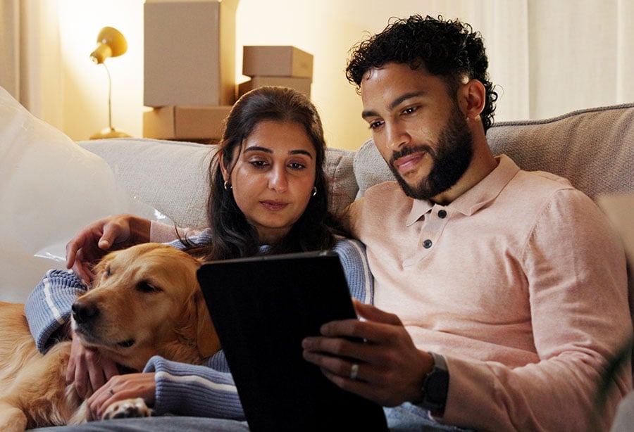 A couple on a sofa look at a tablet, with packing boxes stacked behind them