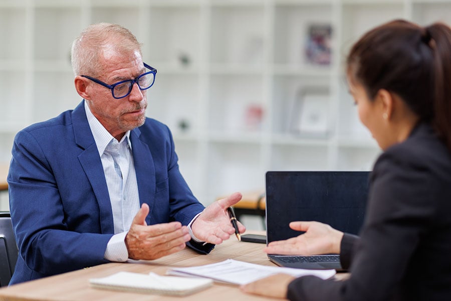 A man and a woman sit on opposite sides of a desk in conversation, with a pile of papers and a laptop between them