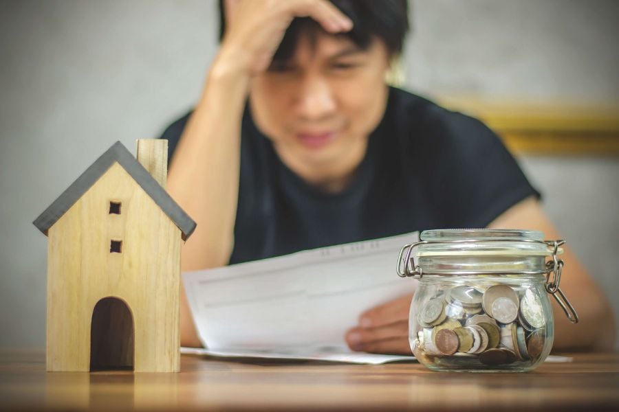 A man looks stressed as he looks at a document. He leans on a table where there are positioned a small wooden house and a change jar full of coins