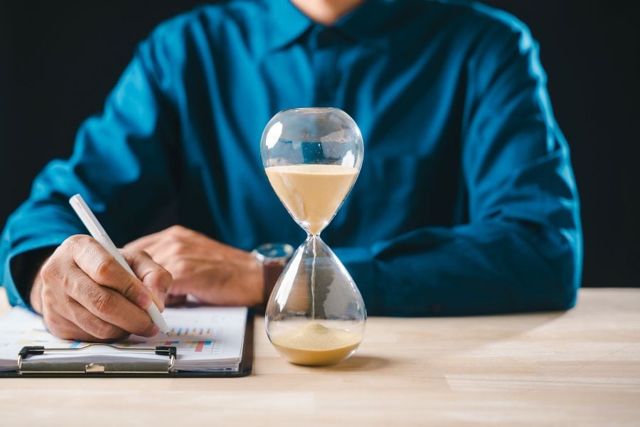 Sand trickles down through an hourglass, which sits on a desk next to a man who is making notes with a pen on paper.