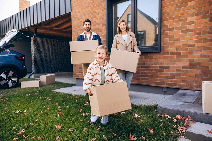 A smiling family with a young child stand outside a house while holding boxes, surrounded by fallen leaves on the lawn