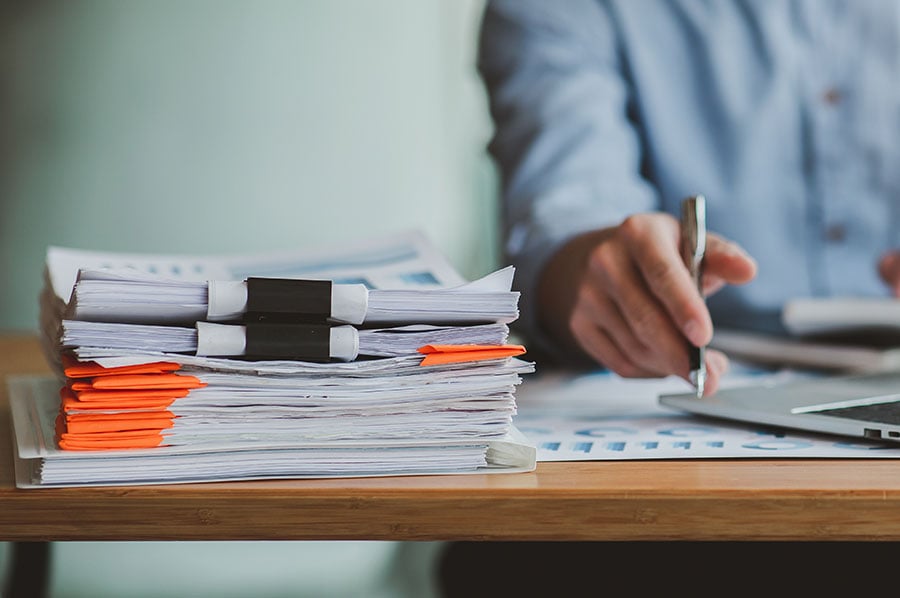 A pile of papers on a desk in front of a man holding a pen