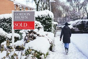 A pedestrian walks down a snowy street, past a house with a sold sign