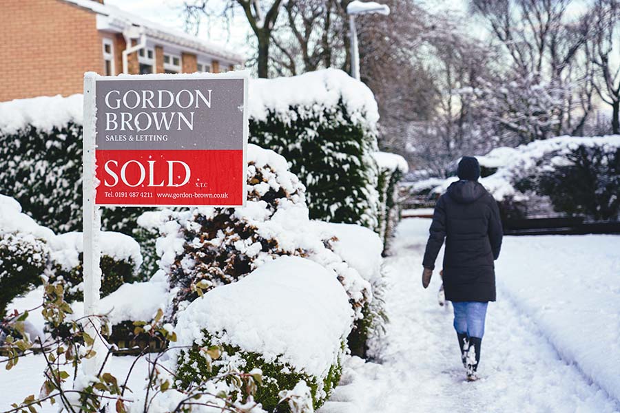 A pedestrian walks down a snowy street, past a house with a sold sign