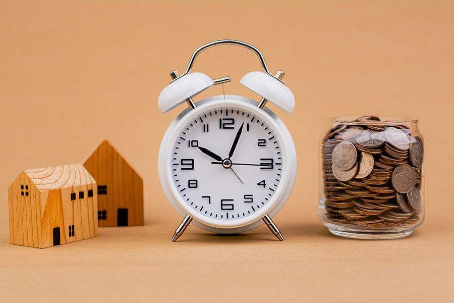An alarm clock next to a small wooden house and a jar full of coins