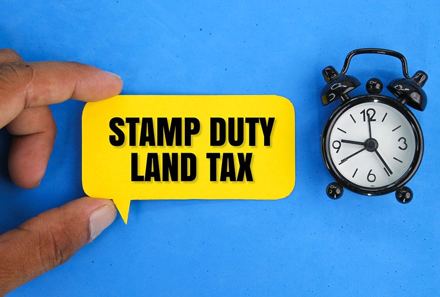 A hand holds a sign with the word stamp duty land tax, with an alarm clock next to it