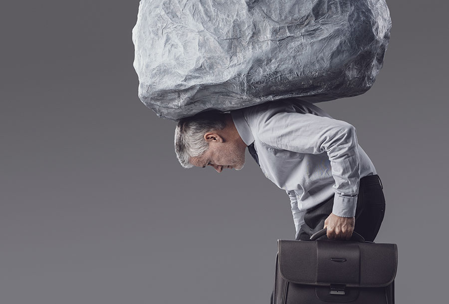A man carrying a briefcase, bent over with a rock balanced on his back
