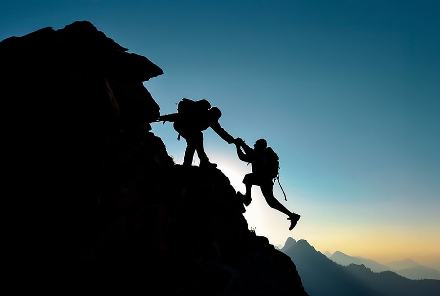 A silhouette image of hikers on a mountain, with one pulling the other up a steep incline