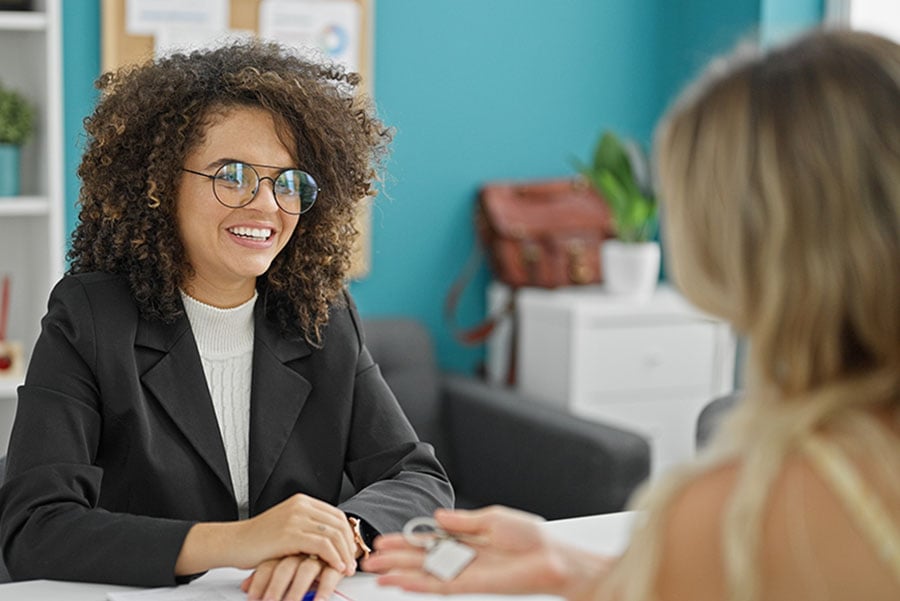 A smiling woman behind a desk talks to a client seen from behind