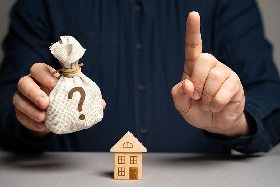 A hand holds a sack with a question mark on it, with the other hand holding up a finger as a warning. A small wooden house is in the foreground